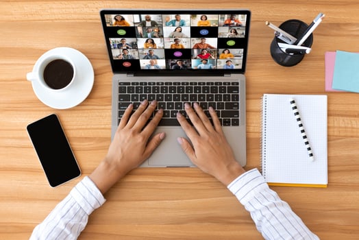 A person is sitting at a wooden desk and using a laptop to participate in a conference. The laptops screen shows a grid of tiles, each displaying a different participant, top view photo