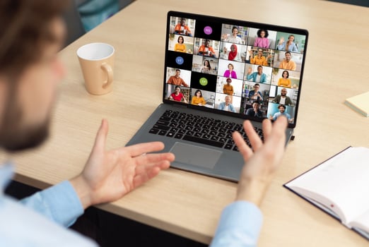 A man sits at a desk in front of a laptop, actively participating in a conference meeting with colleagues. His hands are raised, gesturing and engaging with the online participants. photo