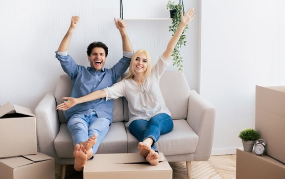 A couple sits on a couch with their arms raised, smiling and celebrating after moving into their new home. photo