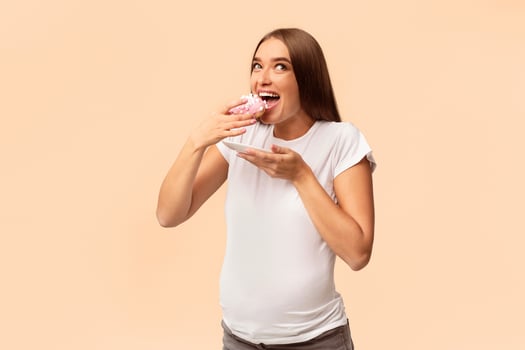 Pregnancy Nutrition. Excited Pregnant Lady Eating Unhealthy Donut Dessert Standing Over White Studio Background. photo