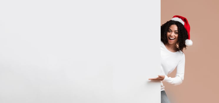 Cheerful Interracial Couple In Santa Hats Holding And Indicating At White Advertisement Board For Your Text, Yellow Background photo
