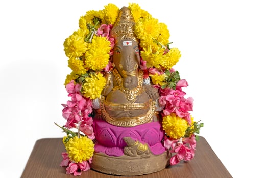 Ganesha statue made of clay decorated with flower garlands on wooden table at home on Ganesha Chaturthi festival celebration. Closeup photo