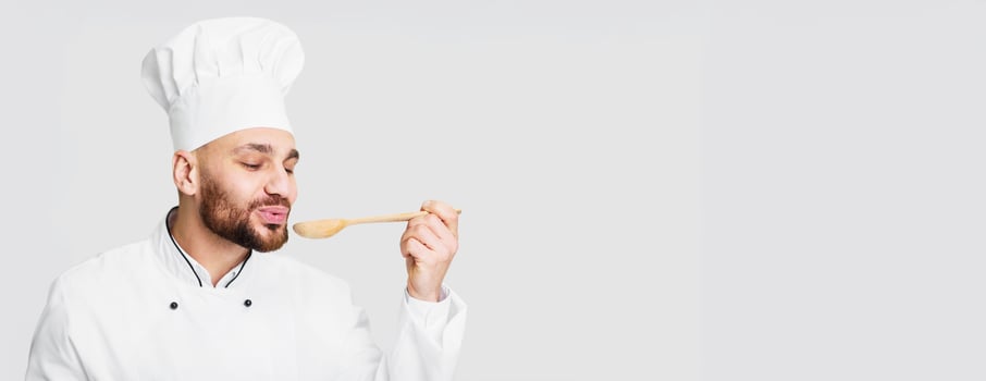 Male Chef Holding Spoon Tasting Delicious Dish Cooking Food Standing Over Gray Studio Background. Panorama, Free Space photo