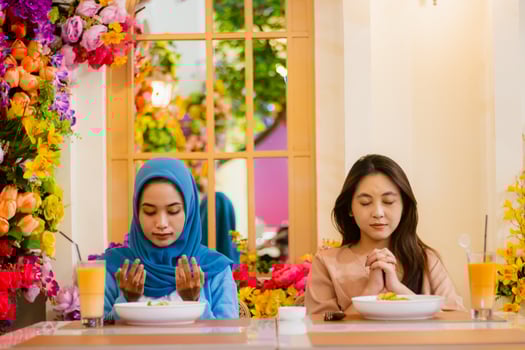 diversity of a pair of beautiful young asian women praying before eating sitting on chairs in an indoor restaurant. for the concept of religion, lifestyle, worship, advertising photo