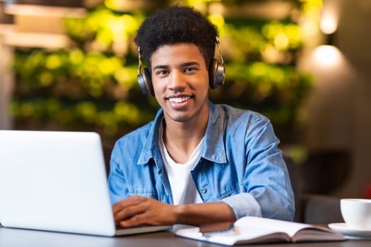 A young black guy student wearing headphones is sitting in front of a laptop, focused on his screen. He appears to be working or studying intently, with a serious expression on his face. photo