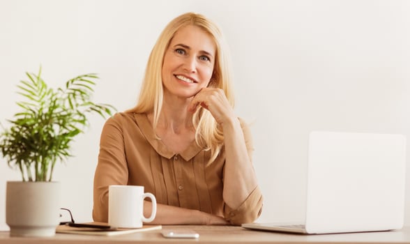 A smiling woman with blonde hair sits at a desk working on a laptop. photo