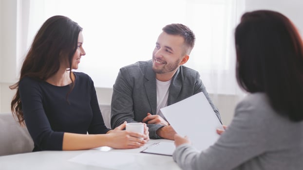 Couple sits at a table in an office, looking at documents with a financial advisor. Advisor holds up a sheet of paper while the couple looks attentive, suggesting they are in the midst of discussion photo