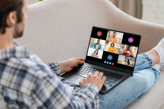 Man sitting on a couch, using a laptop to attend a conference call. The laptop screen displays several participants in a grid format, indicating that it is an online meeting. photo