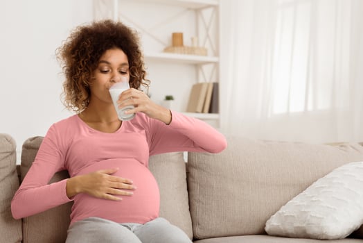Useful diet for pregnant. Afro expectant woman drinking milk, resting on couch, free space photo