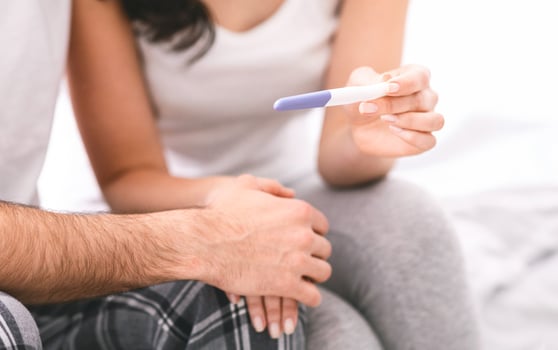 A woman shows a pregnancy test to her partner in bed, with a positive result indicated. The couple is sitting up in bed, and the woman is holding the test up in front of her partner, cropped photo