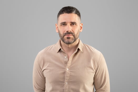 A man with a short haircut and beard is standing against a plain gray background, wearing a beige shirt and looking directly at the camera. The setting appears to be indoors with even lighting. photo