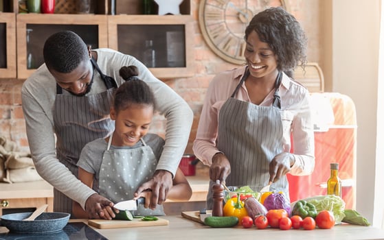 Domestic bliss. African dad teaching daughter how to cut vegetables, mom making salad, kitchen interior, free space photo
