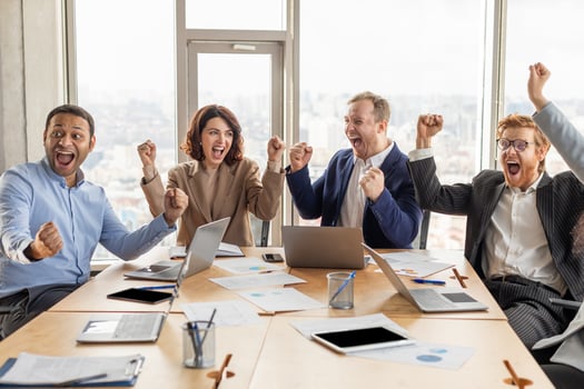 A diverse team of four business professionals are sitting at a conference table in a modern office, all smiling and raising their arms in celebration. photo