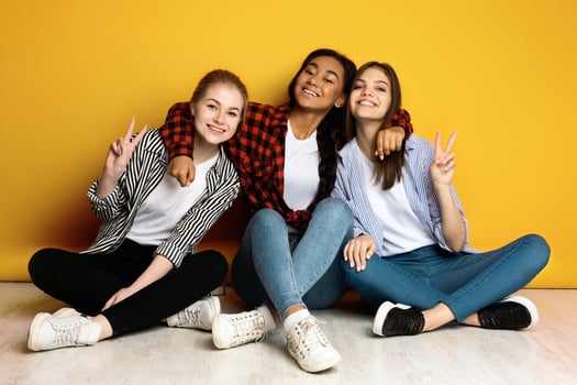 Three young girls are sitting on the floor, striking different poses as if they are getting ready for a group photo. They appear cheerful and excited, forming various poses with their arms and legs. photo