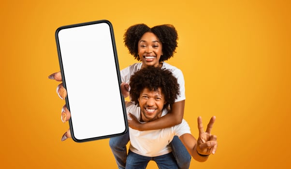 A happy black couple is holding a smartphone with a blank screen in front of a bright yellow background. The woman is giving a peace sign with her right hand and is smiling brightly. photo