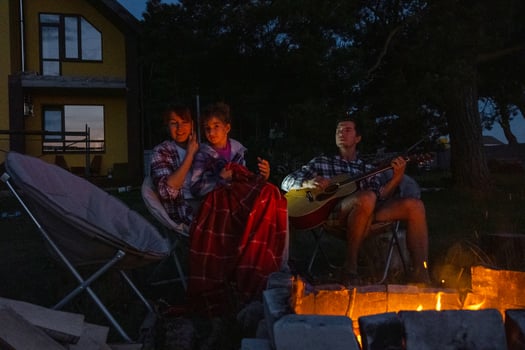 The family is sitting around the campfire, man plays the guitar, a woman listens and sings along, my daughter wrapped herself in a blanket. Family summer evening in the courtyard of the house photo