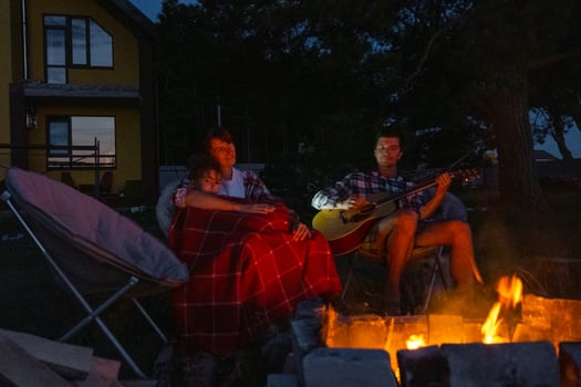 The family is sitting around the campfire, man plays the guitar, a woman listens and sings along, my daughter wrapped herself in a blanket. Family summer evening in the courtyard of the house photo