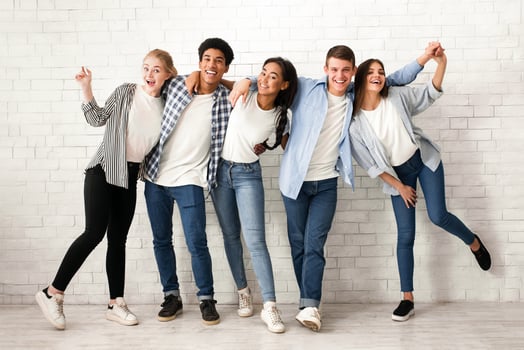 A group of five multiethnic teenagers are standing in a row against a white brick wall, smiling and laughing. They are dressed in casual clothing photo