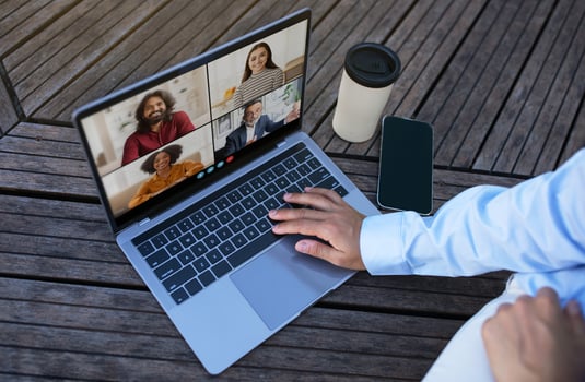 A person sits outdoors on a wooden patio, using a laptop to participate in a conference. They are wearing a light blue shirt and white pants, top view photo