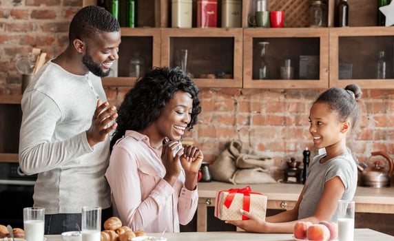 Cute black girl holding birthday gift for her surprised mom, daddy standing behind, kitchen interior, copy space photo