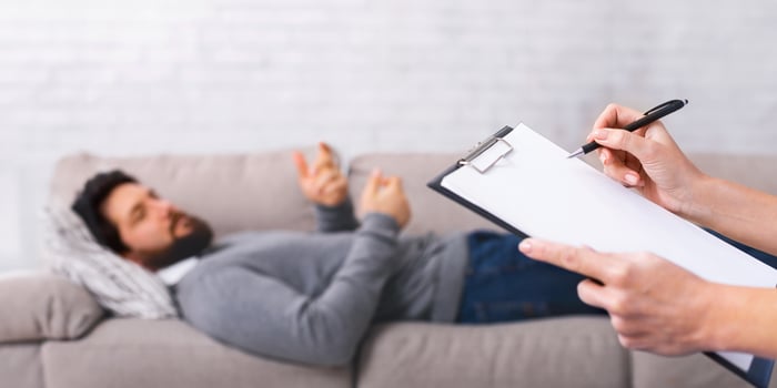 A young man lies comfortably on a gray couch, gesturing with his hands while speaking to someone off-camera. The listener holds a clipboard and pen, ready to take notes or provide feedback. photo