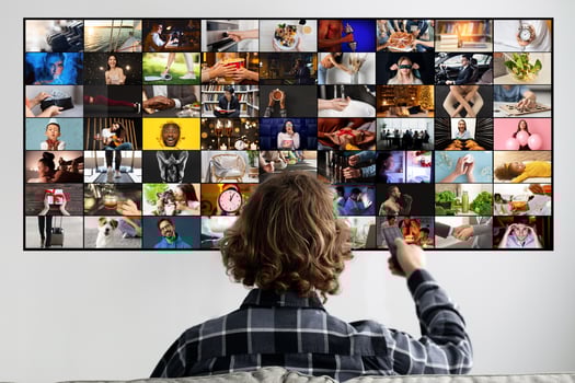 This image shows a man sitting in front of a large television screen. The television is displaying a grid of multiple small videos. The man is holding a remote control in his right hand photo