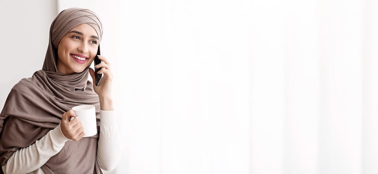 Happy Muslim Girl In Hijab Talking On Cellphone And Drinking Coffee Near Window At Home, Enjoying View, Copy Space photo