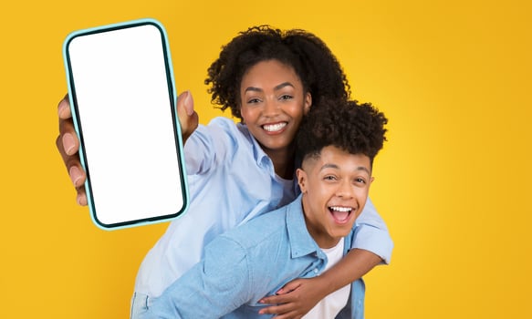 A young black couple, a man and a woman, are standing against a yellow background. The woman is holding a smartphone with a blank screen in front of her, piggybacking her boyfriend photo