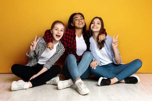 Three young multiethnic girls sit closely side by side with their backs against a bright yellow wall. They are casually dressed in jeans and flannel shirts and are smiling joyfully photo