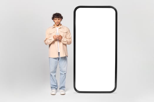 A young man in casual clothing stands in front of a large blank screen, using his smartphone. The screen is large and blank, allowing for space to add your own text or graphics. photo