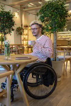Thoughtful dreamy disabled man in wheelchair drinking coffee at cafe table, waiting for meeting photo