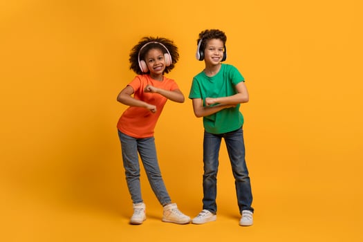 African American young boy and girl stand side by side, each striking a playful pose with their arms while dancing, using wireless headphones, listening to music photo
