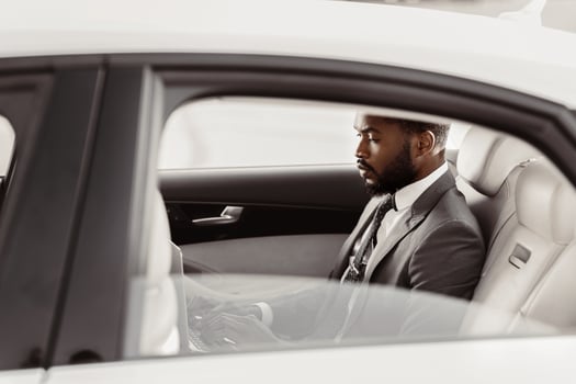 Black man in a suit is sitting in the back seat of a white car. He is looking down and appears to be thinking or working on something. The car is parked in a bright location, likely outdoors. photo