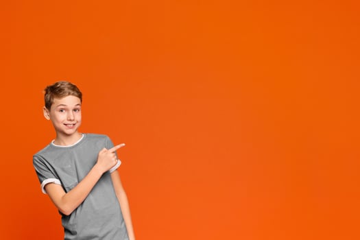 Look over here. Teenage boy smiling and pointing aside at free space on orange background, panorama photo