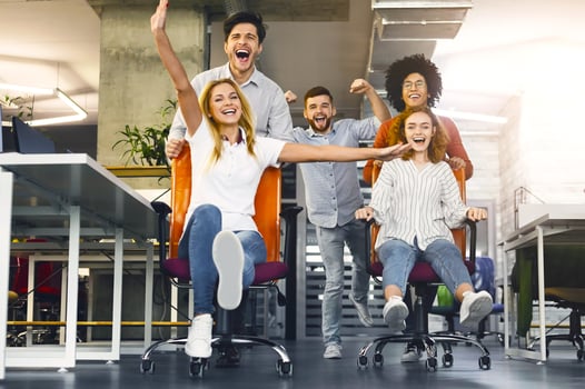 A group of young office workers is caught in a moment of spontaneous joy, racing each other in office chairs. Their expressions are filled with laughter and excitement, suggesting a fun break photo
