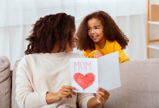Mother's Day Concept. Little Black Girl Congratulating Happy Mom Giving Greeting Card Sitting On Sofa Indoors photo