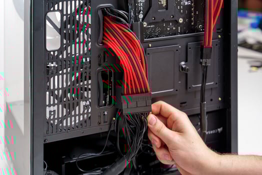 A hand connects a black ATX power supply cable to an Asus motherboard inside a sleek black case. The motherboard features red accents on a black PCB, with a tempered glass side panel for visibility. photo