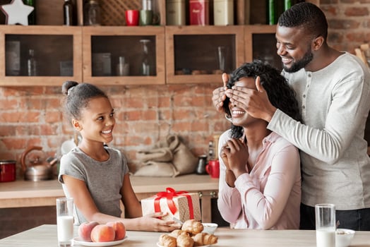 Birthday, mother day concept. Cheerful african little girl holding gift for mother, smiling man closing woman eyes, kitchen interior, free space photo