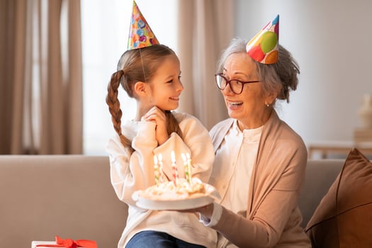 A cheerful grandmother wearing a party hat is presenting a birthday cake with lit candles to a young girl, also in a party hat, as they share a loving glance and a warm photo