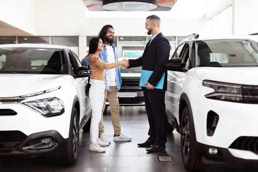 A professional car salesman, dressed in a suit, is shaking hands with a happy Indian couple customers inside a car dealership showroom photo