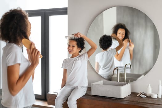 A smiling African American mother stands behind her child, who sits on the counter of a sunlit, modern bathroom, both engaging in a fun hair brushing routine in front of a large circular mirror photo
