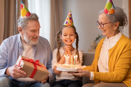 A young girl with a sparkling smile sits between her doting grandparents, all wearing festive party hats, grandmother is holding birthday cake aglow with lit candles, grandfather presents gift photo