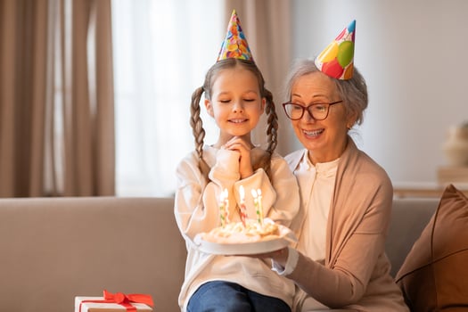 An older woman and a young girl sit on a couch, with a birthday cake in front of them. The woman is lighting the candles on the cake while the girl eagerly watches photo