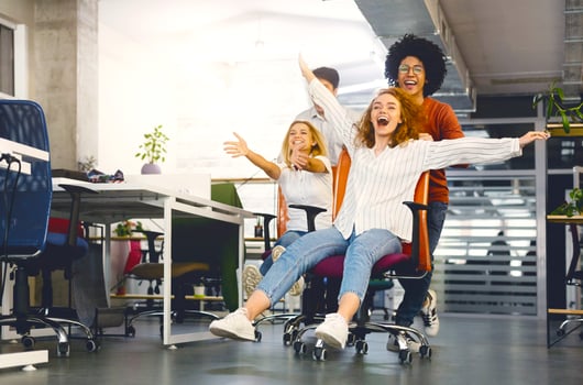 Office fun. Four young cheerful coworkers enjoying office chair race at workspace, empty space photo