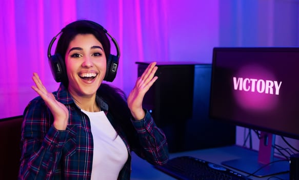 A young woman wearing headphones sits at a computer desk in a dimly lit room, illuminated by pink and blue lights. She is smiling and raising her hands in the air, celebrating her victory. photo