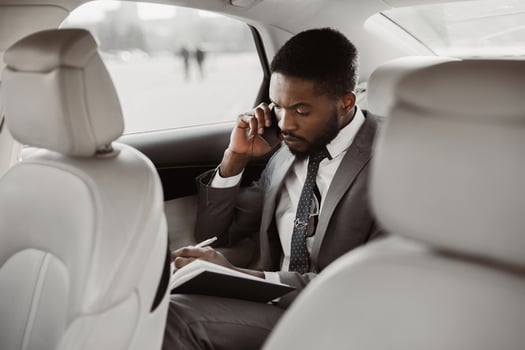 Black man in a suit sits in the backseat of a car, taking notes in a notebook while on a phone call. He is focused and serious, likely discussing important business matters. photo