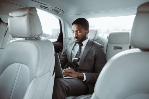 Black businessman in a suit is sitting in the back seat of a car, working on his laptop computer. He is focused on his work, typing on the keyboard. The cars interior is clean and modern photo