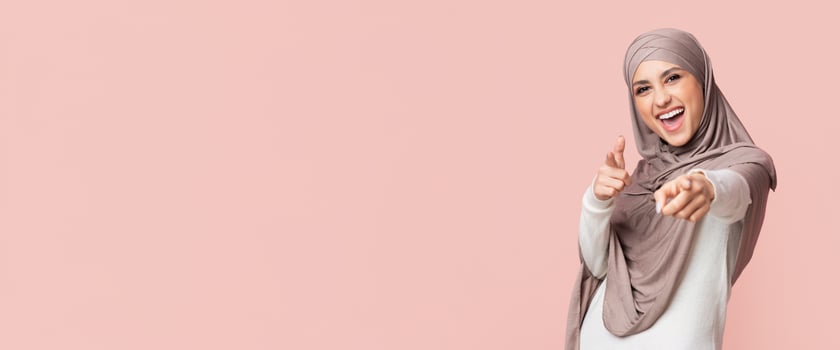 Gotcha. Positive Muslim Girl In Hijab Pointing Fingers At Camera, Posing On Pink Background, Empty Space photo