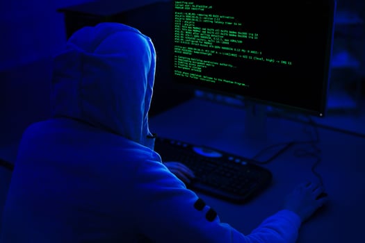 A person wearing a hooded sweatshirt sits in front of a computer with a dark background. Computer screen displays green code, indicating that the person is working on a programming or technical task. photo