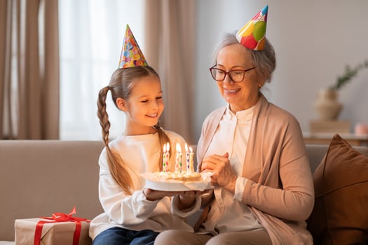 An older woman and a young girl sit on a couch, smiling at a birthday cake in front of them. The woman is lighting candles on the cake while the girl eagerly awaits a slice. photo